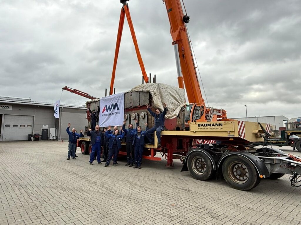 Ein großer Schiffsmotor hängt an orangefarbenen Kranseilen auf einem Schwertransport, davor steht eine Gruppe von Arbeitern auf einem Werksgelände.